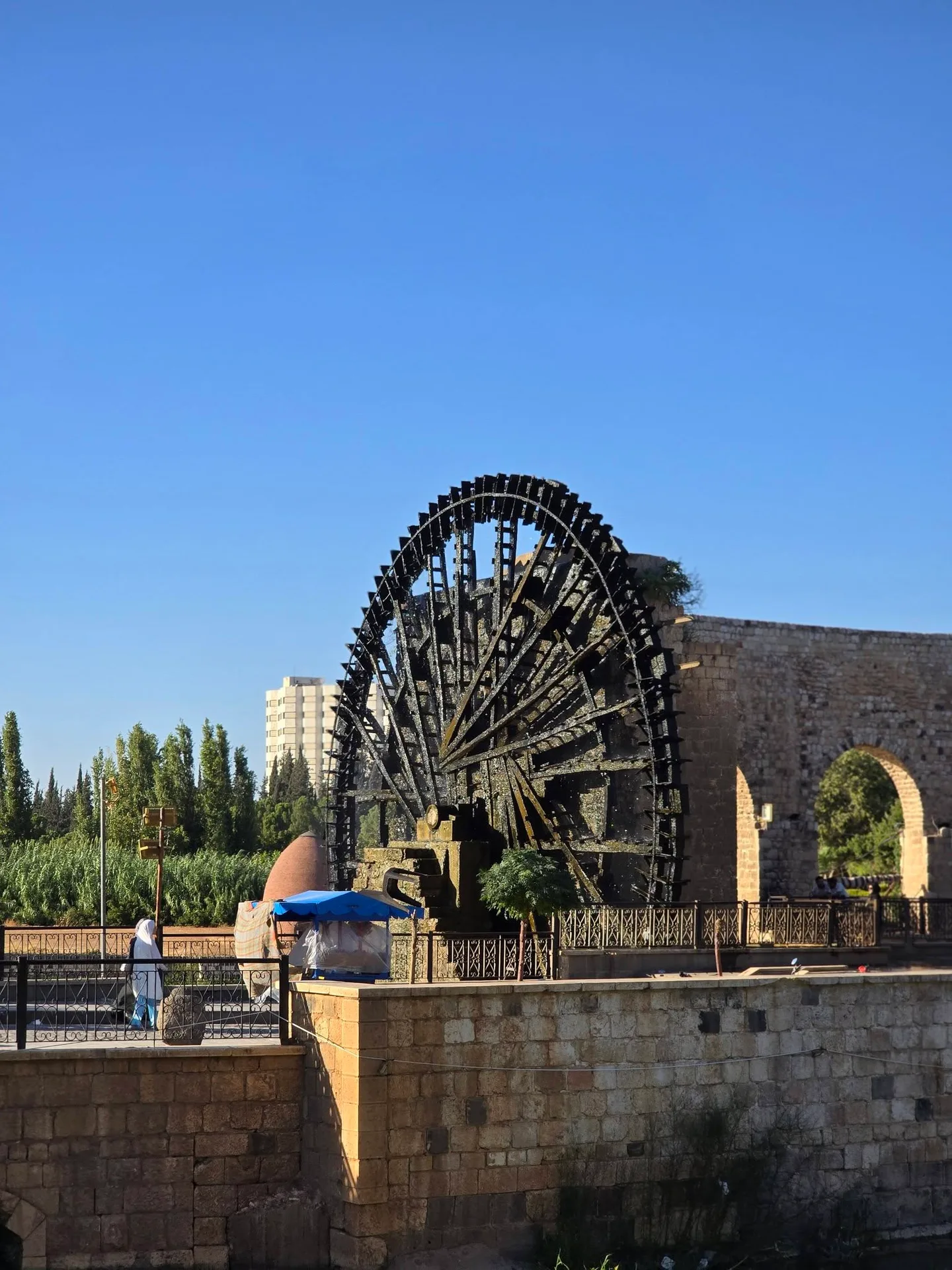 A wooden noria on the Orontes in Hama, the stone aqueduct behind it, a blue stall on the riverbank